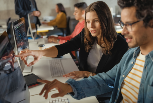 Two people sit at a desk working on computers, collaborating and pointing at screens as they discuss SEO poisoning strategies in an office setting with others in the background.
