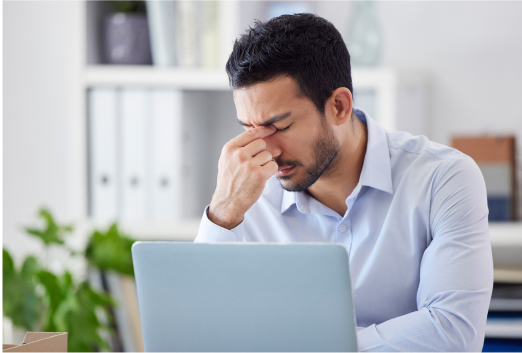 A man sits at a desk in front of a laptop, pinching the bridge of his nose with his eyes closed, appearing stressed or fatigued—perhaps overwhelmed by threats like SEO poisoning.